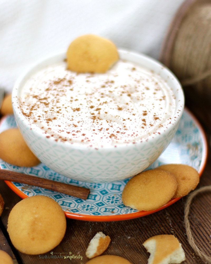 Eggnog dip in a bowl on a plate surround by vanilla wafter cookies and a cinnamon stick. 